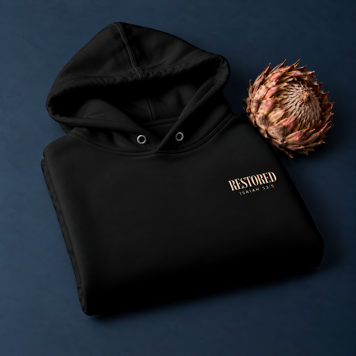 Folded black RESTORED Isaiah 53:5 hoodie beside a dried protea flower